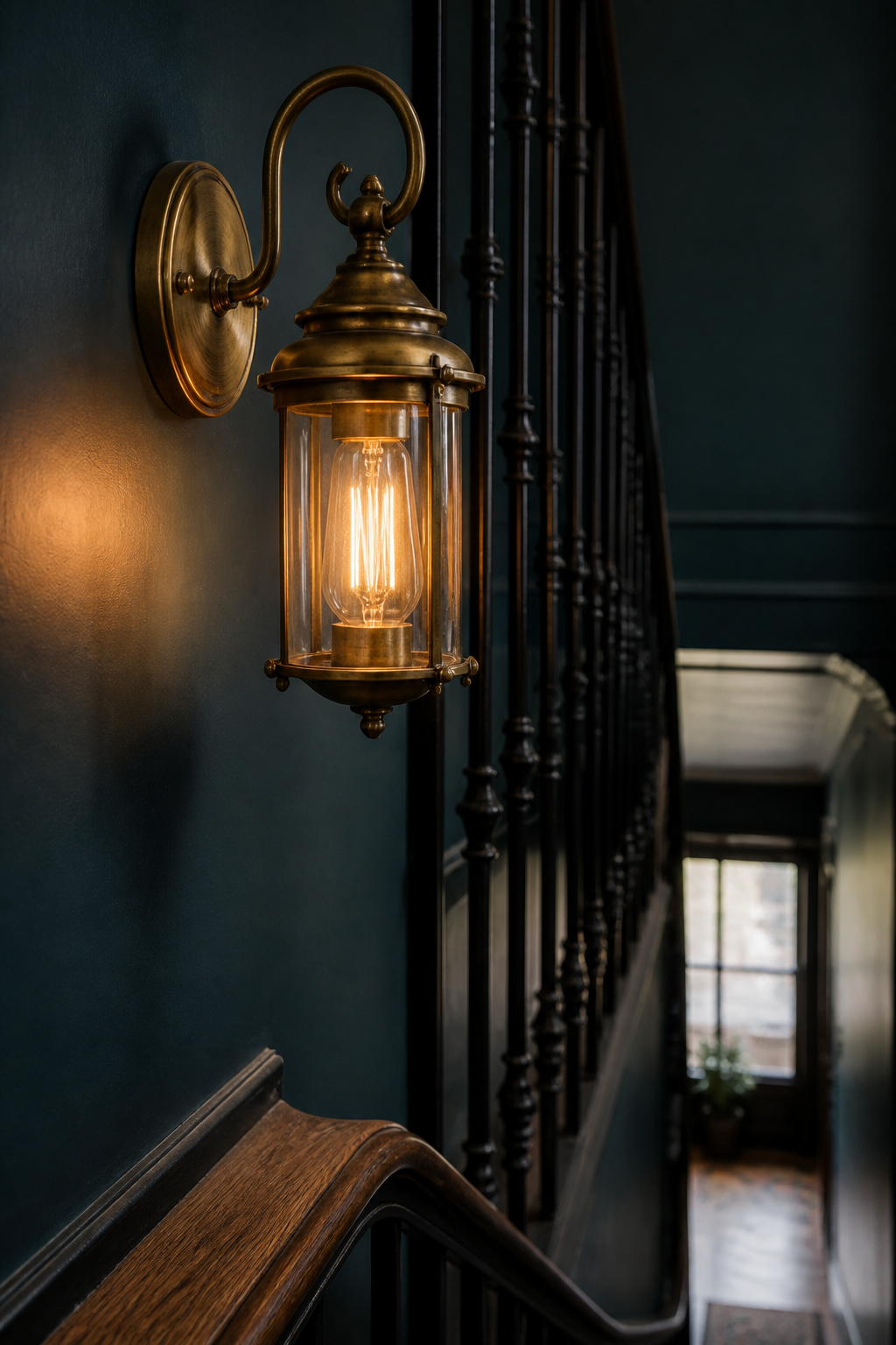 Aged brass wall sconce mounted beside black iron stair balusters and a wood handrail on a deep blue-green stairwell wall
