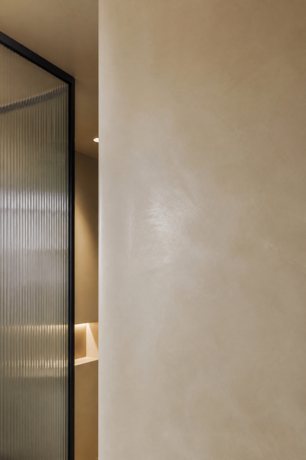 Unbroken bone-coloured microcement wall running floor to ceiling beside a fluted glass shower partition in a small bathroom