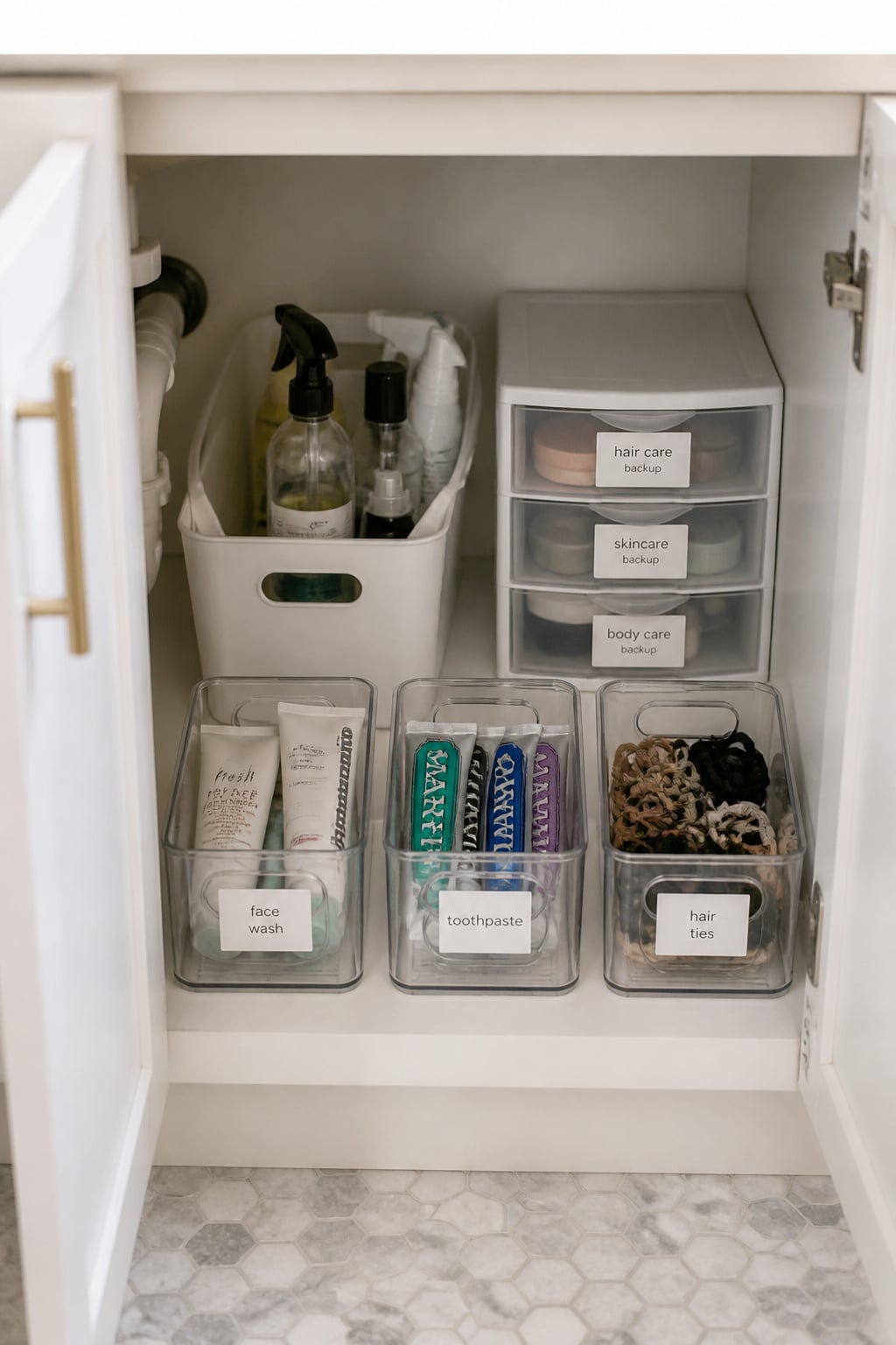 Open bathroom vanity cabinet with clear labelled bins at the front for daily products, white handled cleaning tote at the back, small stacking drawer for backup items