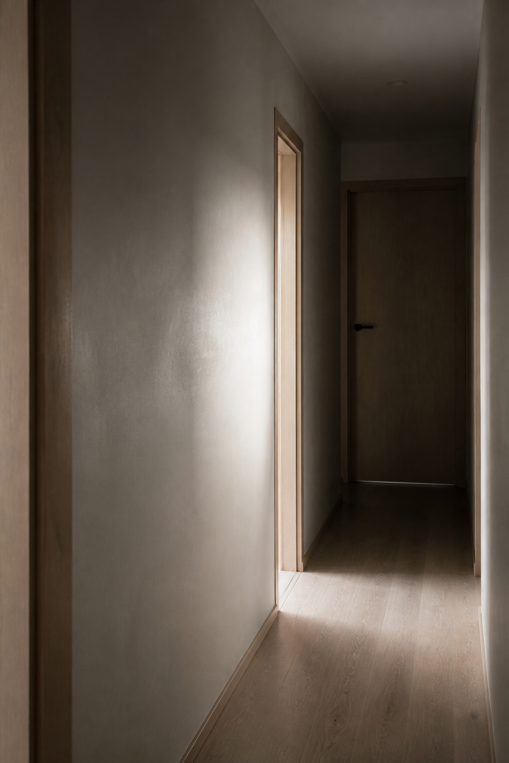 Narrow windowless rental hallway with pale greige walls catching only a sliver of borrowed light from a side doorway, dimming toward a closed door at the far end