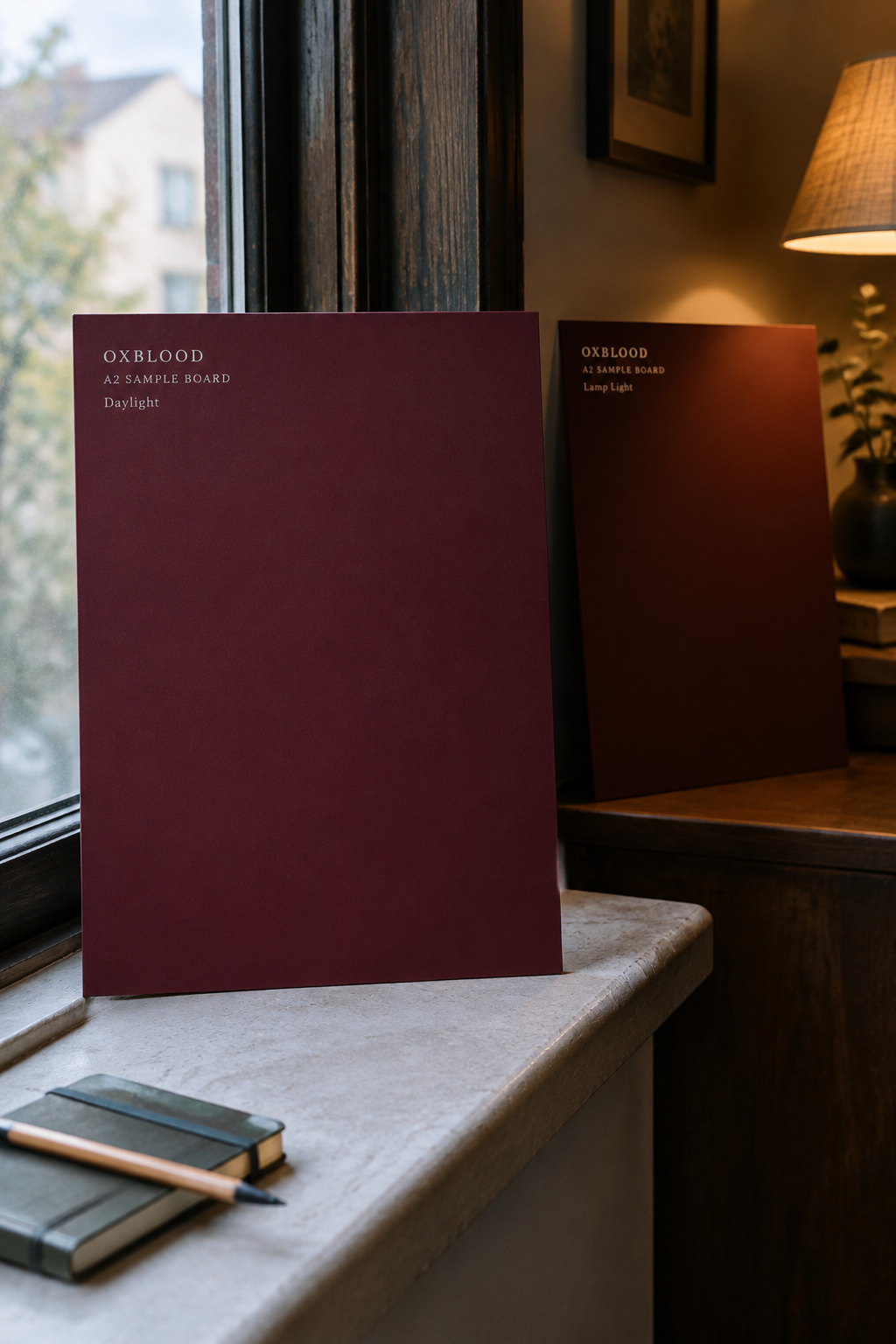 Two matching oxblood sample boards, one in daylight by a window and one in warm lamp shadow, showing the colour shift between light conditions