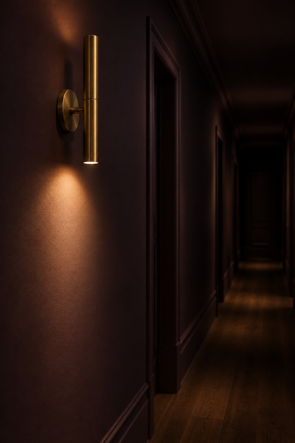 Brass wall sconce mounted at sixty inches casting a warm pool of light on a smoked-aubergine hallway wall, with darkness beyond
