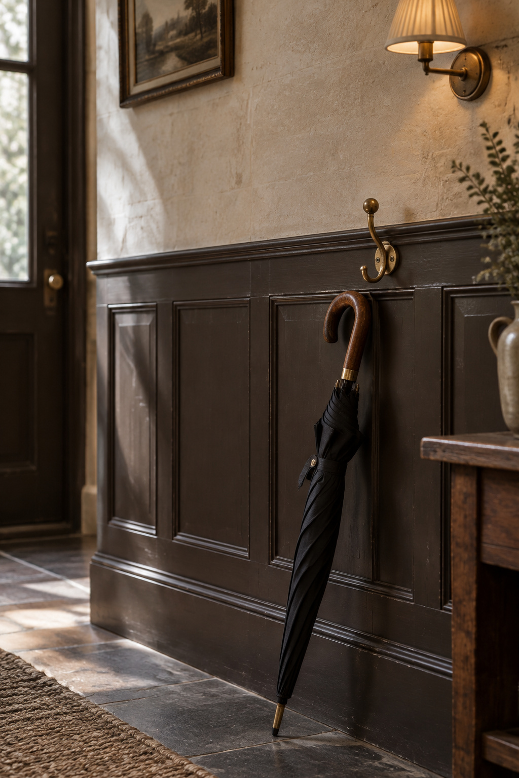 Close view of a dark panelled entryway wall below a dado rail with a brass coat hook and leaning umbrella